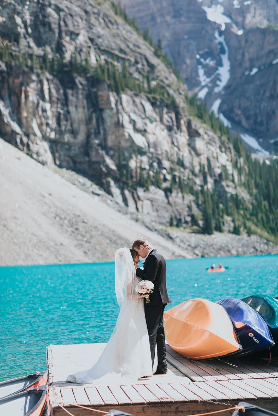 Moraine Lake bride & groom portraits with turquoise blue water, mountains & colorful canoes