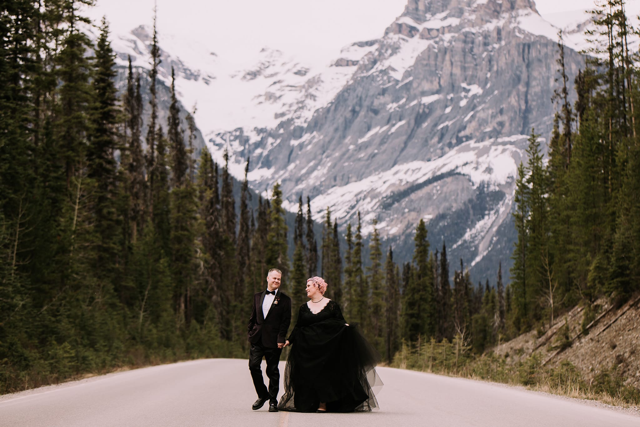 alternative wedding bride black wedding gown groom burgundy tuxedo walking road in mountains