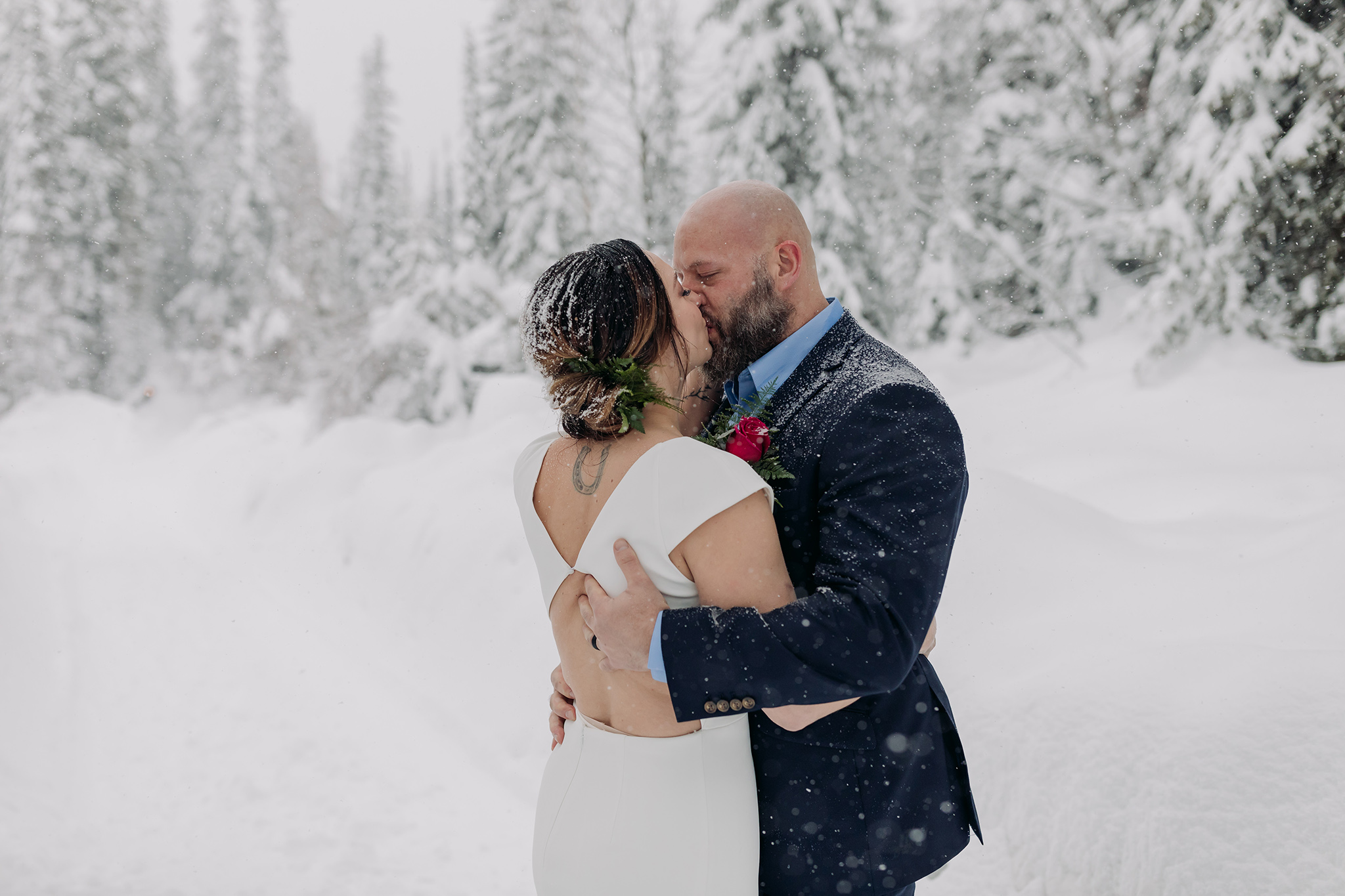 elopement outdoor wedding ceremony first kiss in the pathway amongst the cabin with a natural tree arch with falling snow photographed by mountain elopement & wedding photographer ENV Photography