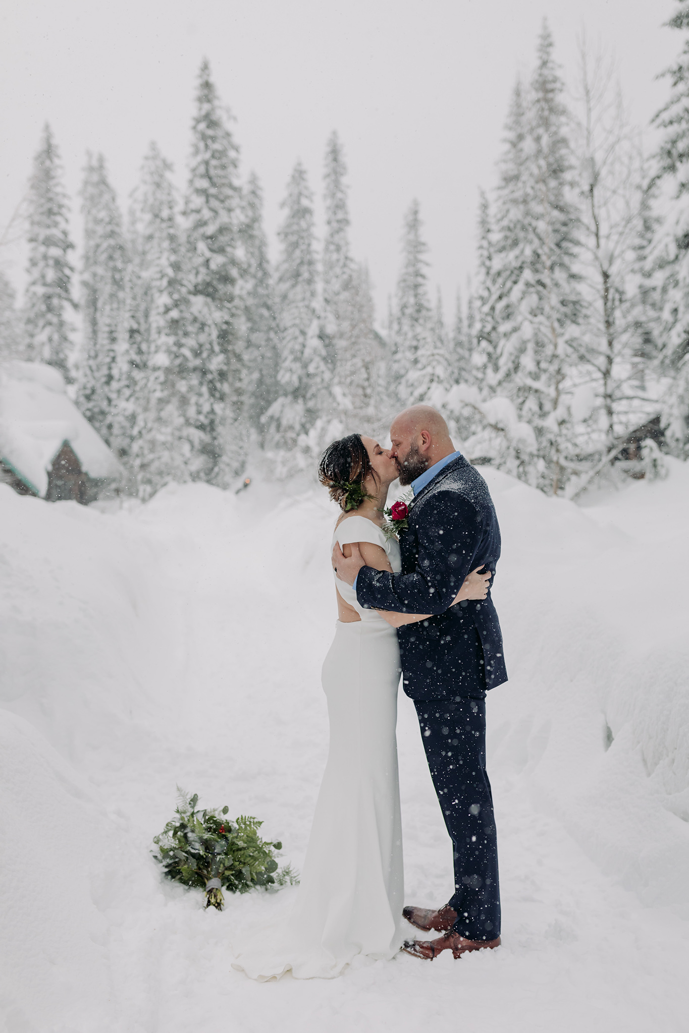 Emerald Lake Lodge winter elopement outdoor wedding ceremony in the pathway amongst the cabin with a natural tree arch with falling snow photographed by mountain elopement & wedding photographer ENV Photography