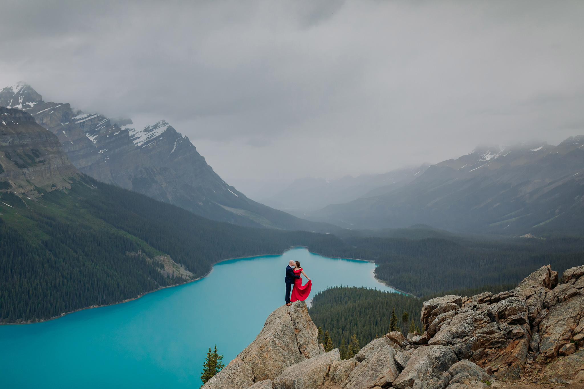 Peyto Lake adventurous Formal engagement photos on a cold rainy day in the mountains in Banff National Park. Featuring stunning red evening gown & dramatic blue lake backdrop photographed by ENV Photography