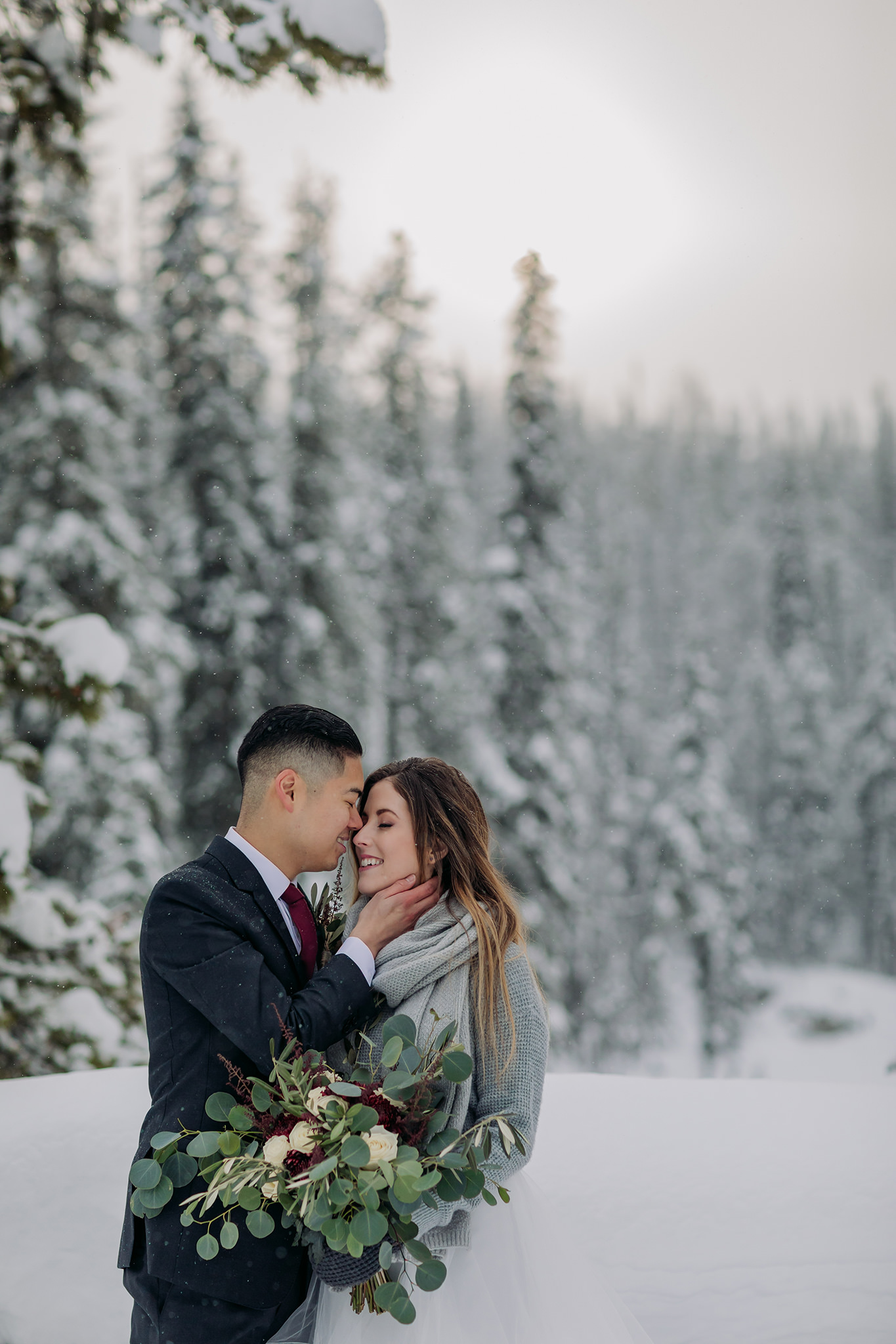 Snowy winter elopement portraits at the natural bridge in Yoho National Park. Mountain wedding in a winter wonderland. Bride in a cozy sweater & matching scarf to keep warm. What to wear for a winter wedding. 