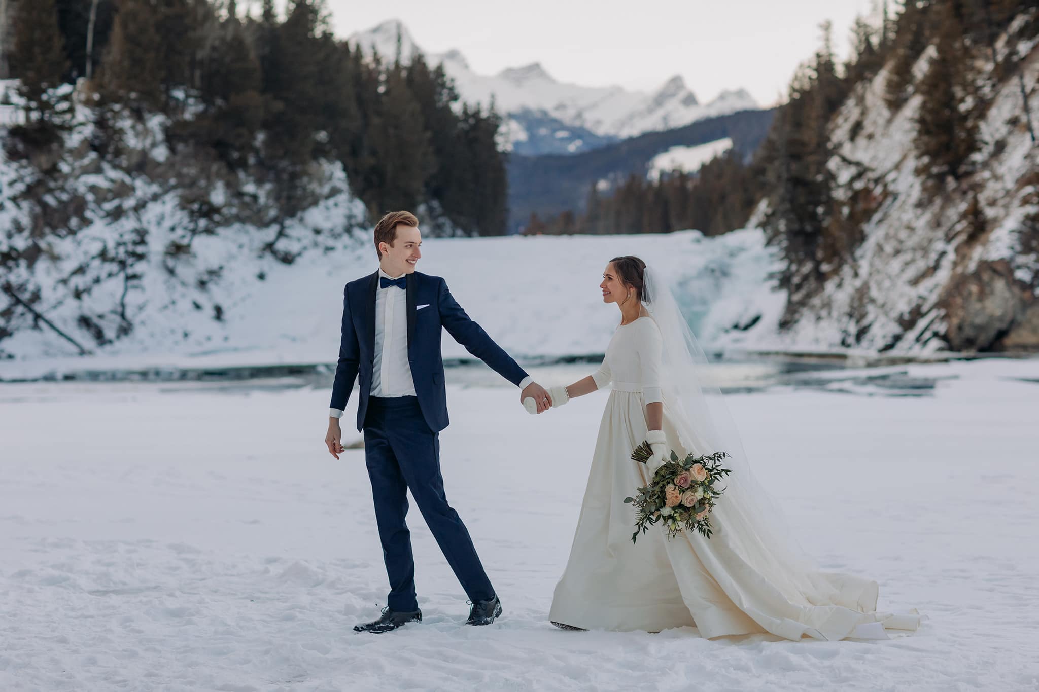winter wedding couple walking on frozen Bow River at Bow Falls in Banff National Park